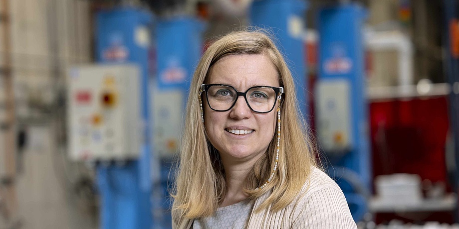 A blonde woman with glasses looks friendly into the camera. Blue machines can be seen in the background. The woman is standing in a workshop.