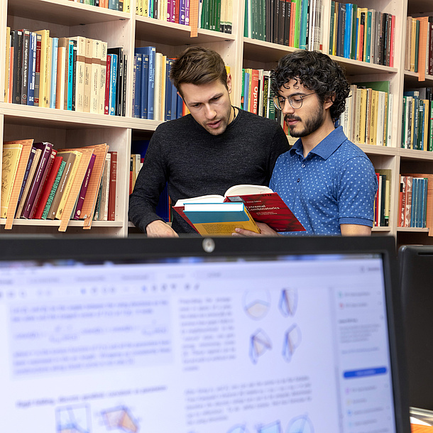 Two men are standing by a bookshelf and looking into a book.