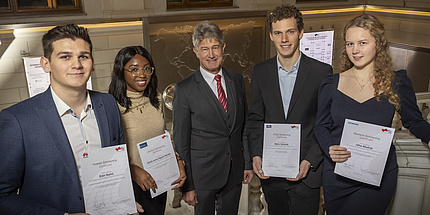 Five people, two women and three men, smile into the camera. Four of them show their certificate which indicates them receiving a scholarship.