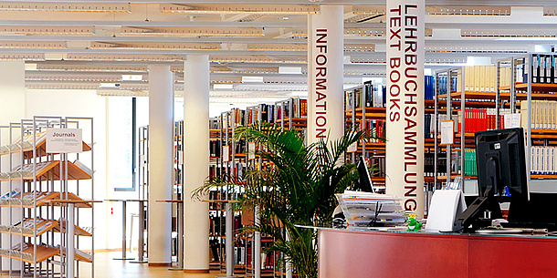 Bookshelves and a service counter with a computer in a large room. One column is marked INFORMATION and another is marked LEHRBUCHSAMMLUNG.