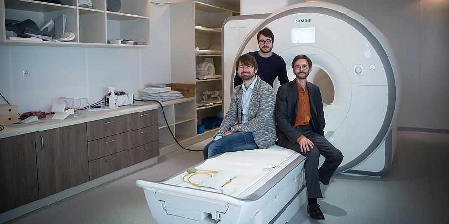 Three gentlemen sit in front of an MRI examination tube on the corresponding couch.