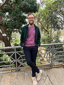 A man leans against a bridge railing and smiles into the camera