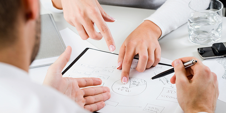 Zwei Personen sitzen sich gegenüber und besprechen ein auf Papier gezeichnetes Kreisdiagramm. Zu sehen sind im Bildausschnitt nur die Hände der Personen und das Diagramm, eine Person hält einen Kugelschreiber in der rechten Hand.