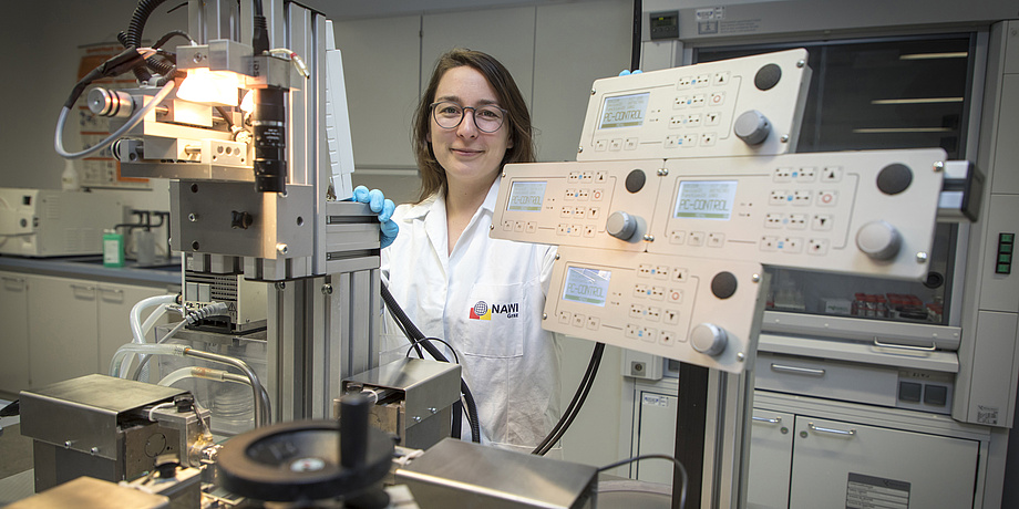 A young woman in a lap coat stands inside a laboratory. Complicated testing equipment surrounds her.