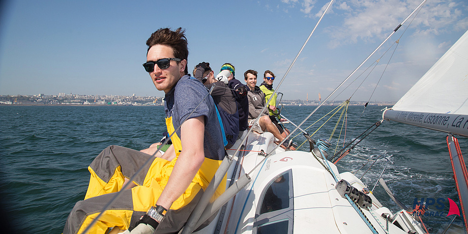 Six young sailors sitting in a row on a sailing boat which is slightly leaning over.