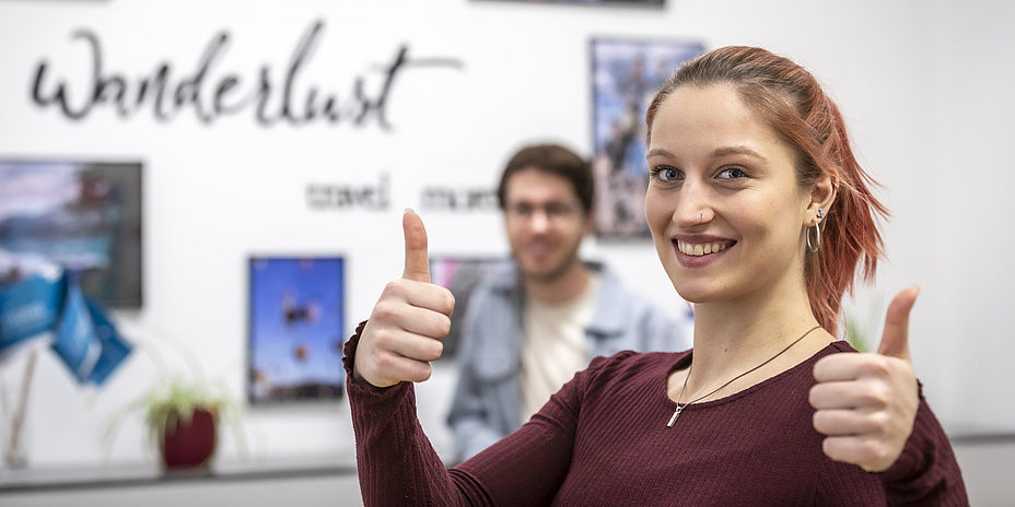 A young woman stands in front of a decorated wall with the inscription "Wanderlust" and gives both thumbs up.