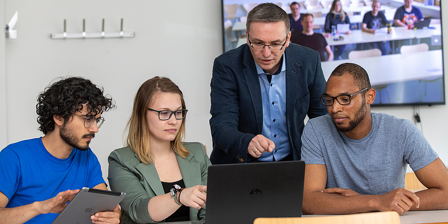 Two young men, one with tablet and young woman sitting in front of a laptop and an older man in gray suit standing behind them pointing to something on the laptop..