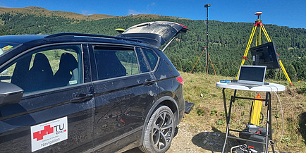 Ein Auto steht neben einem kleinen Tisch, auf dem ein Computer steht. Hinter dem Tisch steht eine GNSS-Antenne.