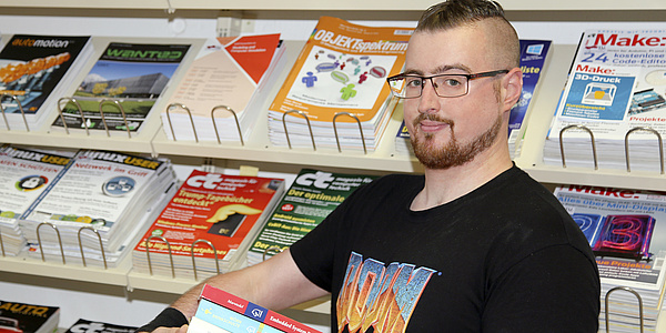 Mann steht vor einem Regal mit Zeitschriften und hält Bücher in der Hand