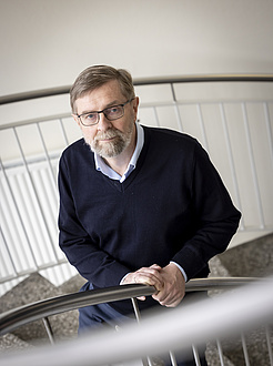 A man with a grey beard and glasses stands at a banister and looks into the camera.