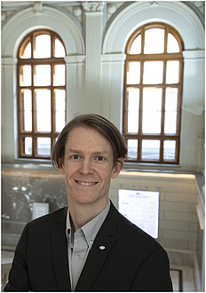 A man in a blue shirt smiles, standing in front of two large windows.