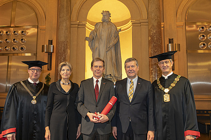 Fünf Menschen stehen vor der Statue von Kaiser Joseph II., die äußeren beiden Herren tragen einen Talar, jener in der Mitte hält eine rote Urkundenrolle in Händen. 