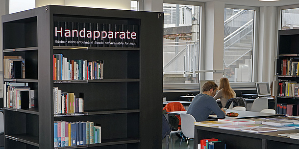 A bookshelf with a sign ‘Handapparate’ In the background, people are sitting at tables with books.