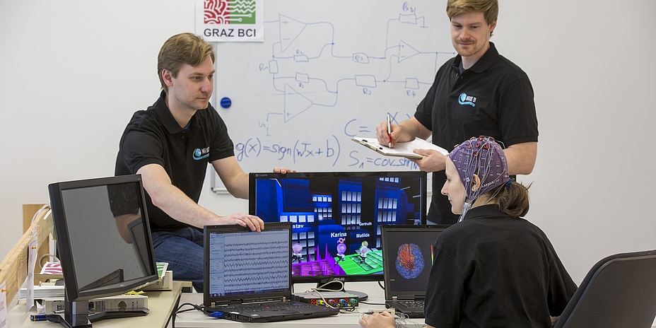 Eine Frau mit Datenhaube sitzt vor mehreren Computerbildschirmen, zwei Kollegen sehen sie an. Im Hintergrund ist eine Tafel mit mathematischen Formeln zu erkennen.