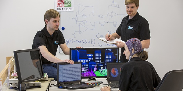 Eine Frau mit Datenhaube sitzt vor mehreren Computerbildschirmen, zwei Kollegen sehen sie an. Im Hintergrund ist eine Tafel mit mathematischen Formeln zu erkennen.