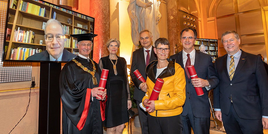 Festively dressed group of people with scrolls of certificates, one person is watching via television