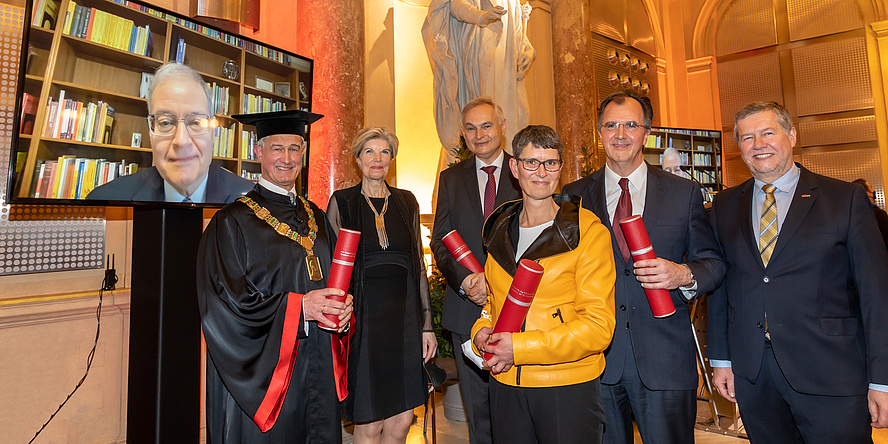 Festively dressed group of people with scrolls of certificates, one person is watching via television