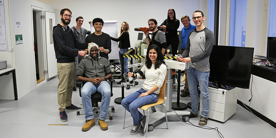 Mitglieder des BCI Racing Teams 2018 mit diversen technischen Assistenzsystemen in einem hell eingerichteten Arbeitsraum am Institut für Neurotechnologie.