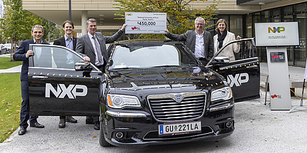 Five people, four men and one woman, stand next to a car and hold up a cheque with a value of 450,000 US dollars