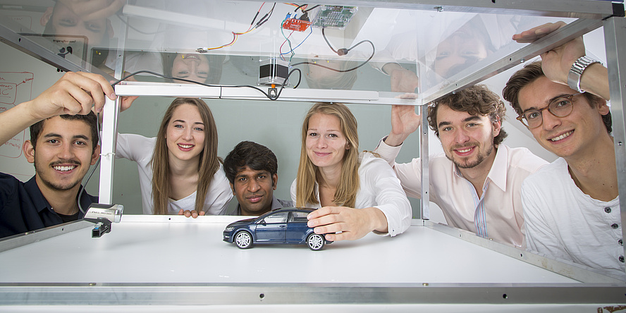 sechs junge Menschen betrachten ein kleines Spielzeugauto in einem Glaskasten