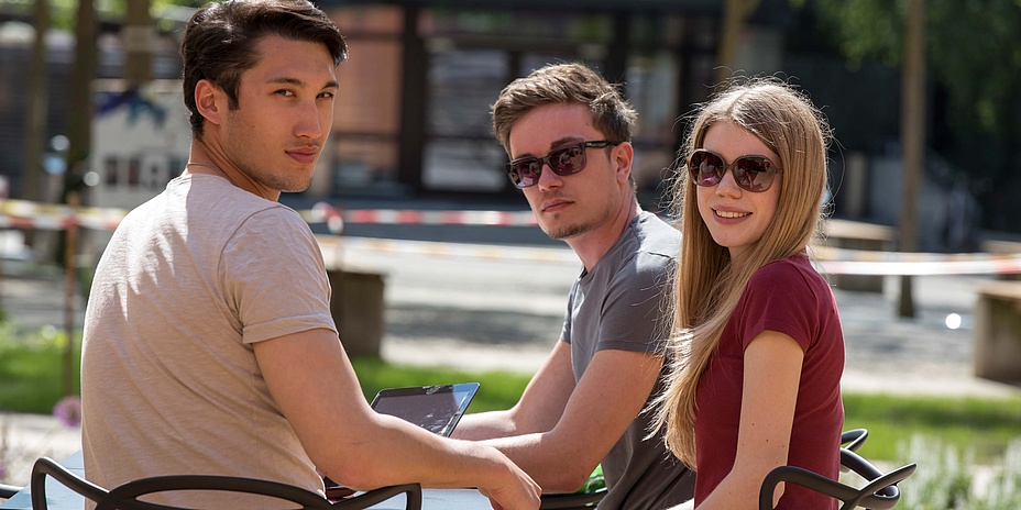 Zwei junge Männer und eine junge Frau im Gastgarten eines Cafés.