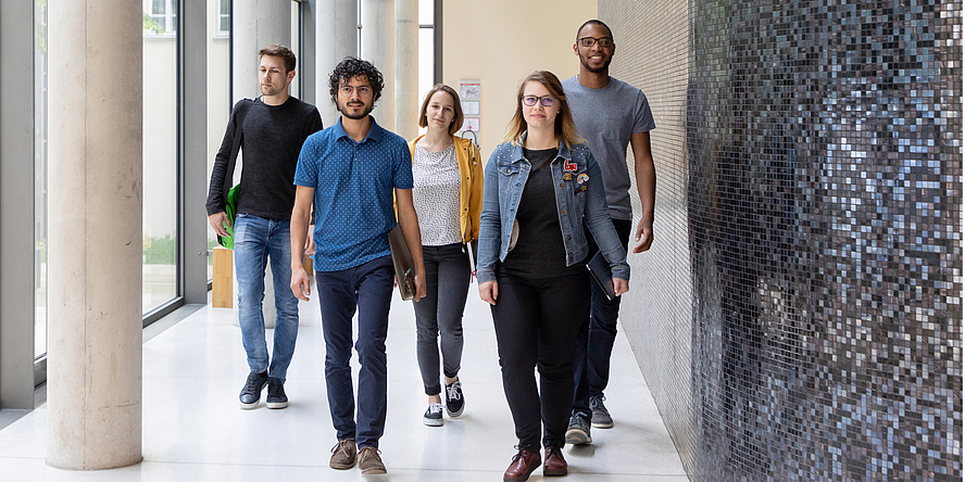 Drei Studenten und zwei Studentinnen gehen mit Schwung einen Gang entlang auf den Betrachter oder die Betrachterin des Fotos zu.