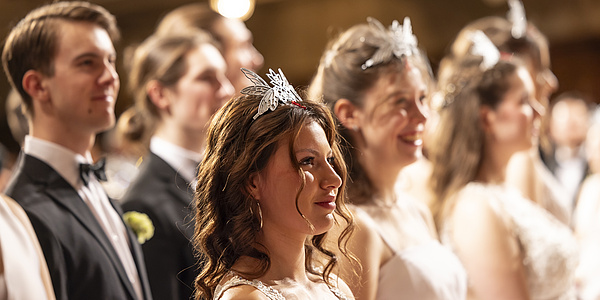 Eine lächelnde Frau mit festlichem Haarschmuck im Vordergrund, umgeben von festlich gekleideten Männern und Frauen.