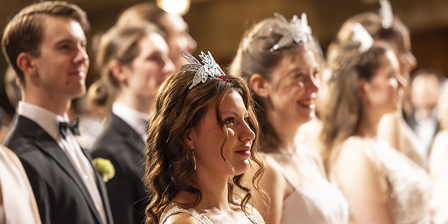 Eine lächelnde Frau mit festlichem Haarschmuck im Vordergrund, umgeben von festlich gekleideten Männern und Frauen.