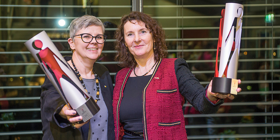 Two women hold up tube-shaped, red-silver trophies and smile into the camera.