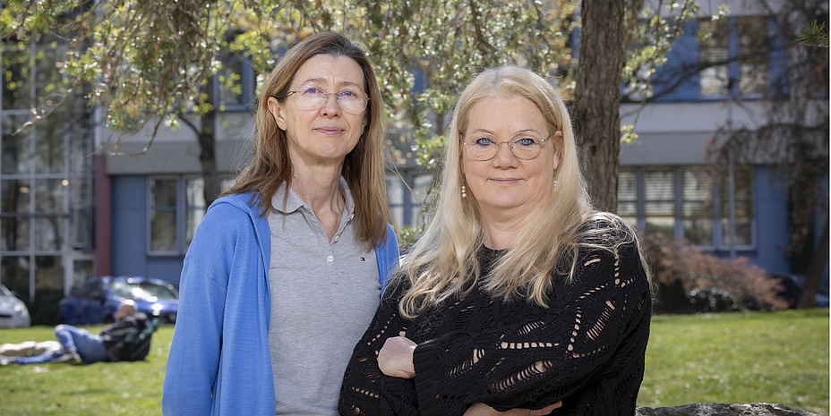 Two women are looking at the camera. One woman is wearing a blue vest and glasses; the other woman has blonde hair and is wearing a black sweater.