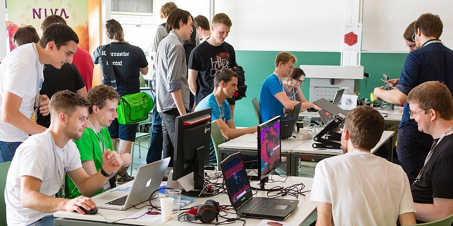 Eine Gruppe Menschen stehen und sitzen um mehrere Tische herum, auf denen Computer und Monitore stehen.