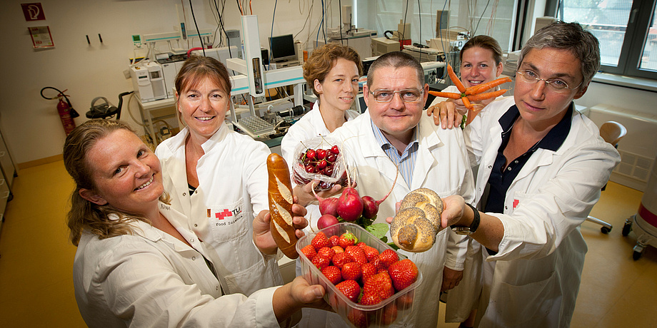 Six people dressed in white laboratory clothing are holding different foods.