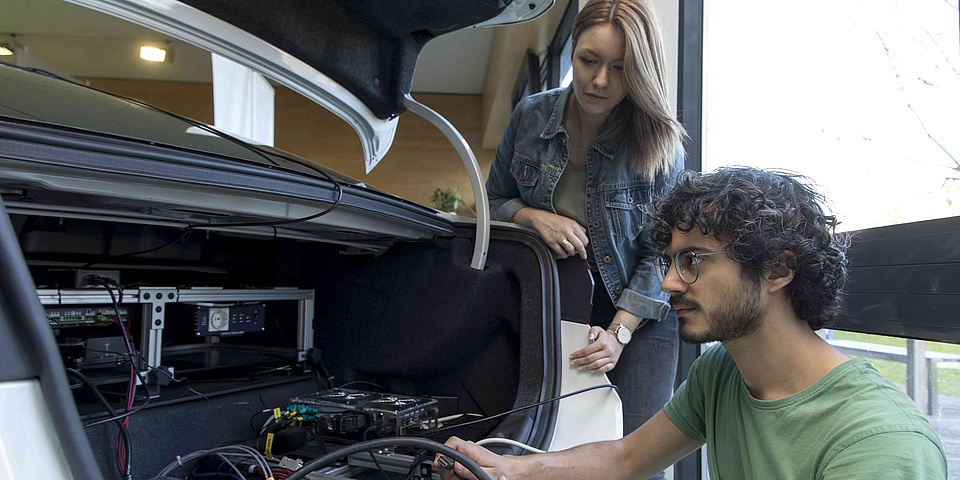 Junge Frau und junger Mann vor dem geöffneten Kofferraum eines Testautos mit technischen Geräten und vielen Kabeln.