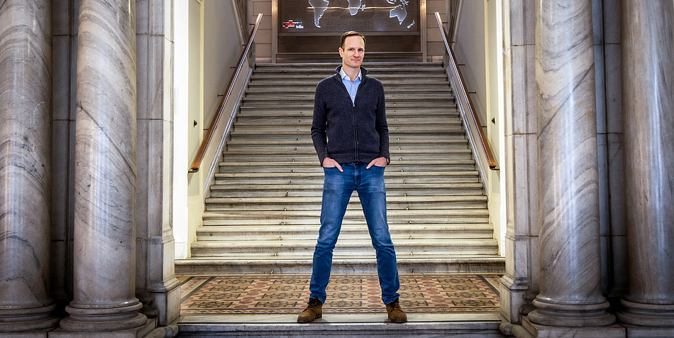 Ein Mann steht breitbeinig vor einem Treppenaufgang. Er hat die Hände in seine Hosentaschen verschränkt und wirkt selbstbewusst. 
