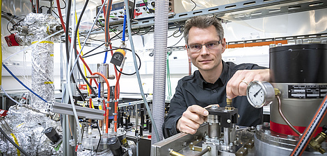 Ein Mann mit Brille steht in einem Labor zwischen mehreren Schläuchen und Metallzylindern.