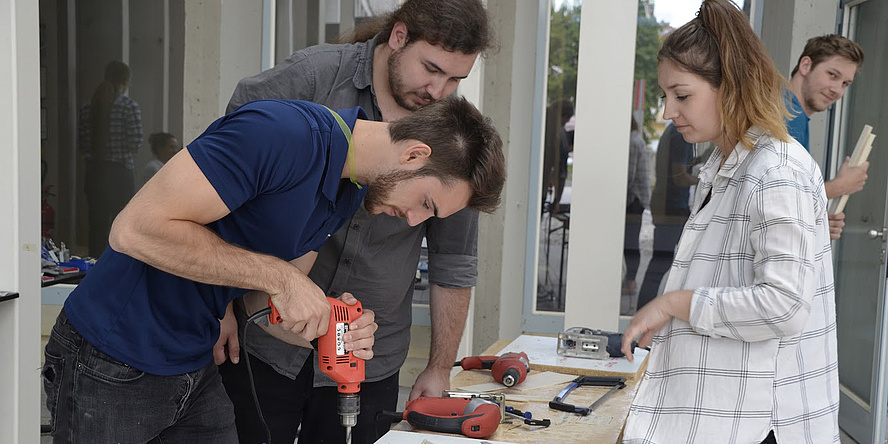 Zwei Studenten und eine Studentin stehen um einen Werktisch und bauen beim Ingenieurswettbewerb EBEC an der TU Graz mit Bohrmaschine und anderen Werkzeugen an einem Prototypen.