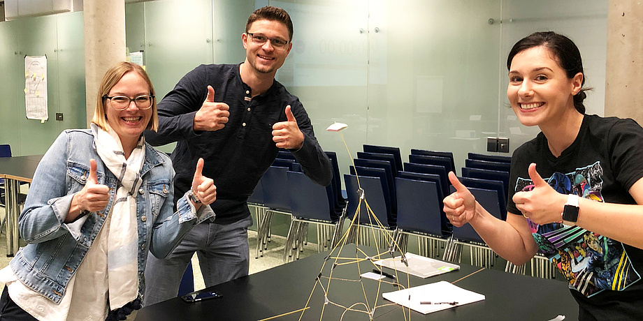 Zwei Frauen und ein junger Mann freuenn sich über den errichteten freistehenden Spaghetti-Turm mit einem Marshmallow an der Spitze.