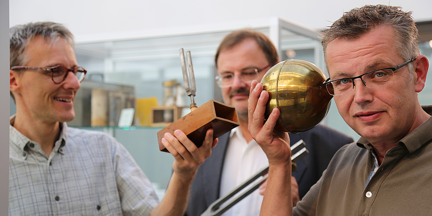 There are three men on the picture. Two of them are holding golden historic tuning forks. The third one on the right is looking at the camera and pointing a golden globe at his ear.
