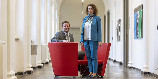 A woman and a man smile at the camera. The woman is standing and the man is sitting on a red armchair to her left.