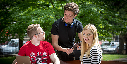 Three young people, a man in a red T-shirt next door, a man in a black T-shirt and a young woman with blond hair sit in nature and exchange ideas.