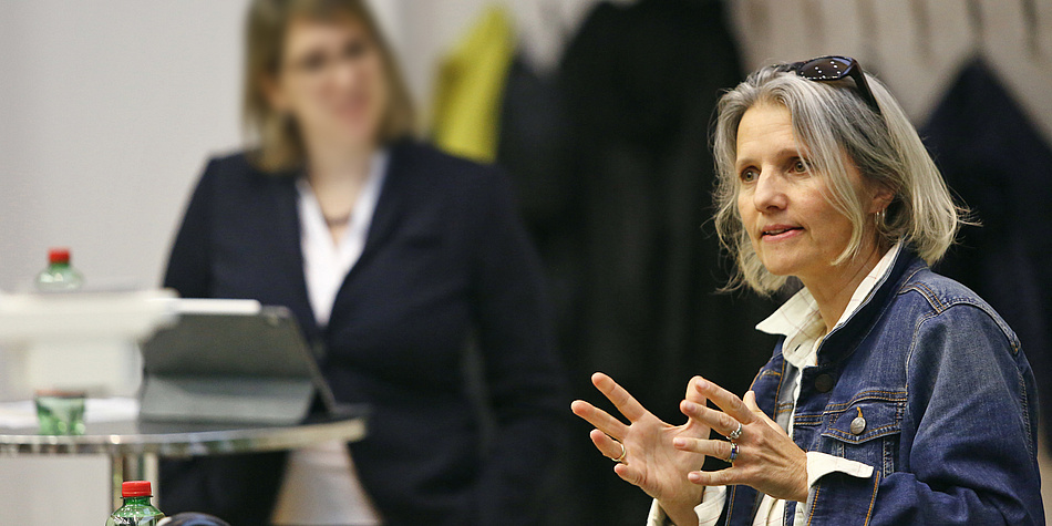 A woman with light hair wearing a blue denim jacket is talking and gesticulating with her hands. Another woman stands in the background and listens.