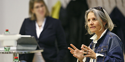 Eine Frau mit hellen Haaren, die eine blaue Jeansjacke trägt, spricht und gestikuliert mit den Händen. Im Hintergrund steht eine weitere Frau und hört zu.