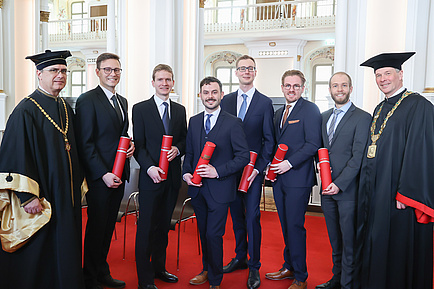 Eight men smile into the camera. The man on the far left and the far right wear festive robes, the six men in the middle hold red document rolls.