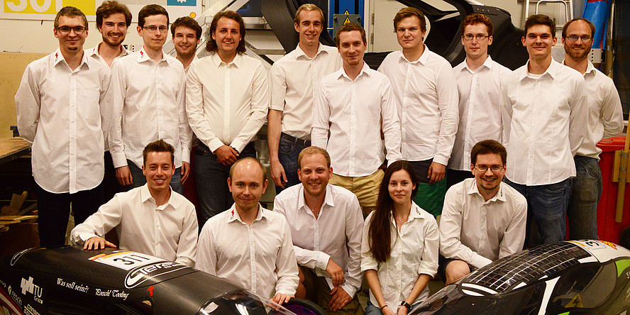 Die Mitglieder des TERA TU Graz Teams – 15 Jungs und ein Mädchen, alle in weißen Blusen – posieren in zwei Reihen für das Foto, im Vordergrund links und rechts sind zwei Elektrofahrzeuge ausschnittweise im Bild.