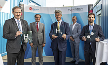 5 men in dark suits look into the camera. Two of them hold a board with the hydrogen sign H2 into the camera. 