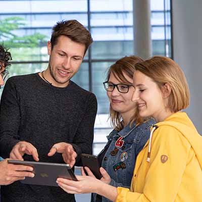 Drei Studierende schauen auf ein Tablet und diskuttieren. 