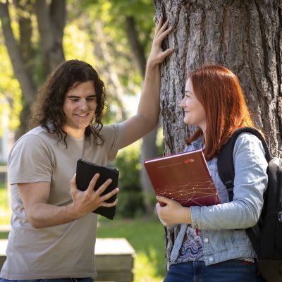 Student lehnt an Baum und zeigt Studentin etwas auf einem Tablet.