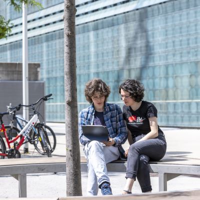 Zwei Studierende sitzen draußen am Campus Neue Technik. Eine Person zeigt der anderen etwas am Laptop.