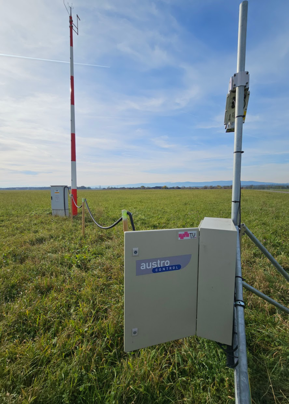 Ein Antennenmast steht auf einer Wiese und ist durch ein Kabel mit einem Matallkasten verbunden, der in einigen Metern Abstand an einem kleineren Mast hängt.
