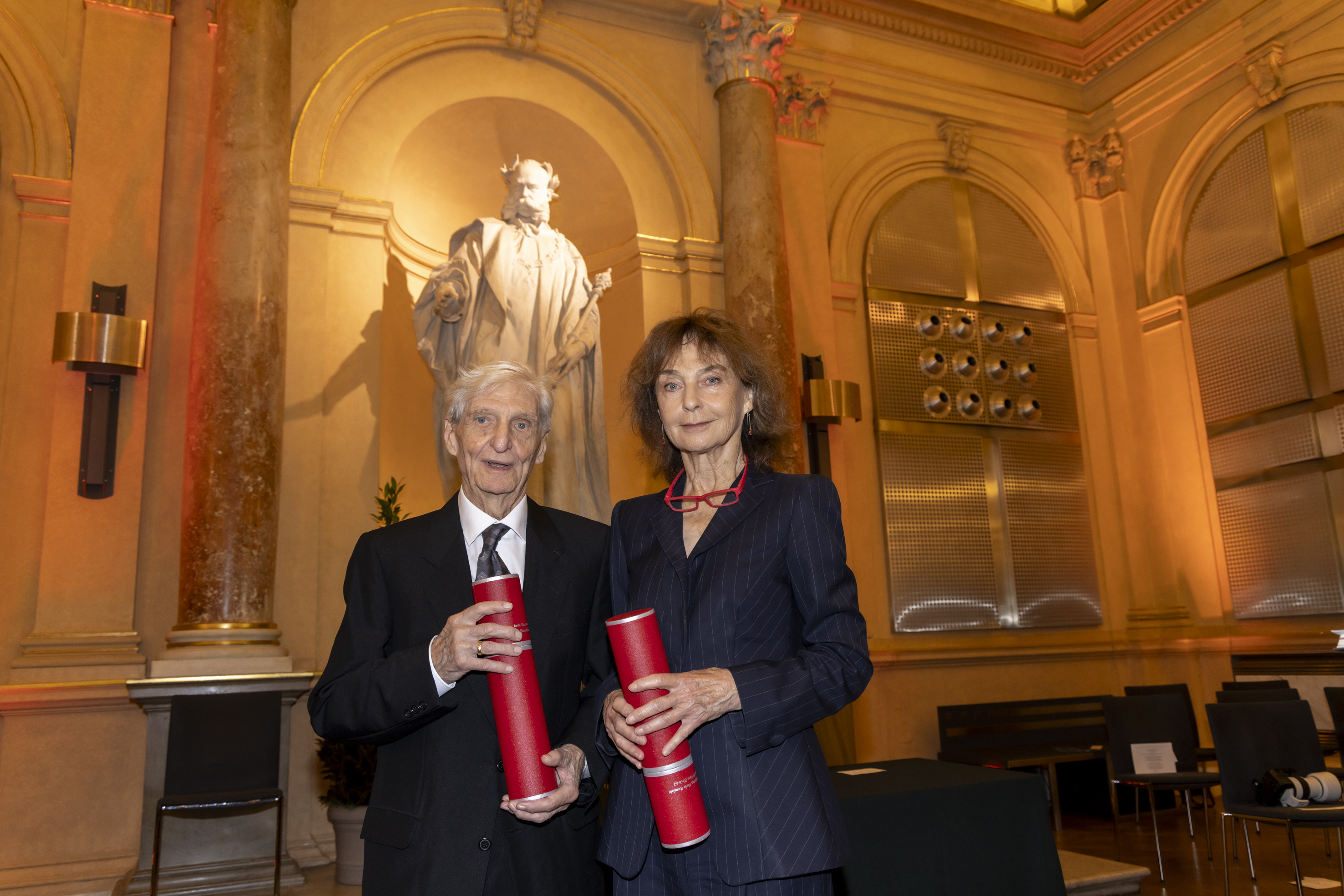 Zwei Personen mit Rollen für Ehrenurkunden in der Hand lächeln in die Kamera. Rechts steht eine Frau, links steht ein Mann.